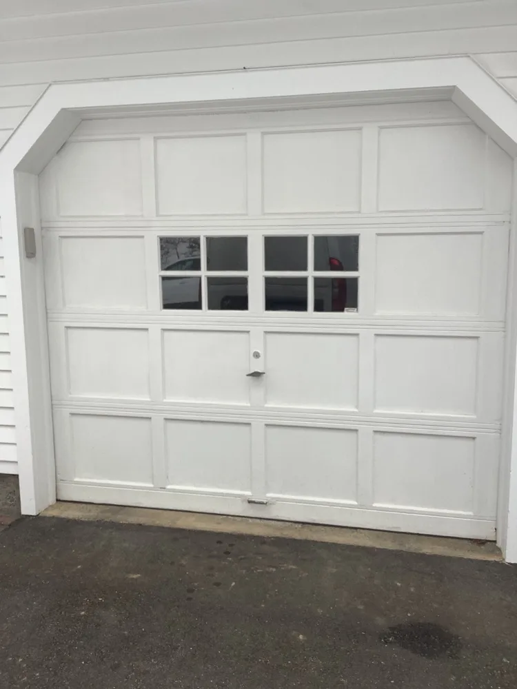 A white raised-panel garage door with an eight-lite window row across its middle section, set under an arched white trim head on a clapboard garage.