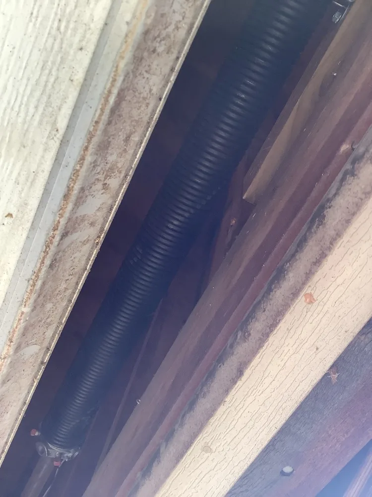 A torsion spring running along its shaft in the tight gap between the top of a garage door and the timber framing above it, the surfaces dusty and unpainted.