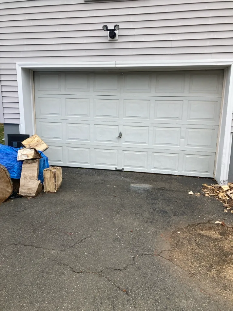 A wide white raised-panel garage door on a clapboard house with a floodlight above it, seen across a cracked asphalt driveway with split firewood stacked to one side.