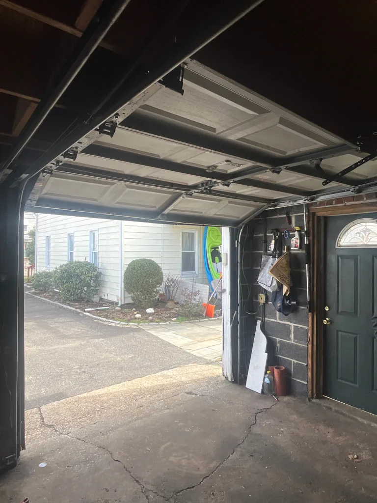 A garage door part way up its curved track, the lower sections still vertical and the upper ones turning into the horizontal run, seen from inside a garage looking out to the driveway.