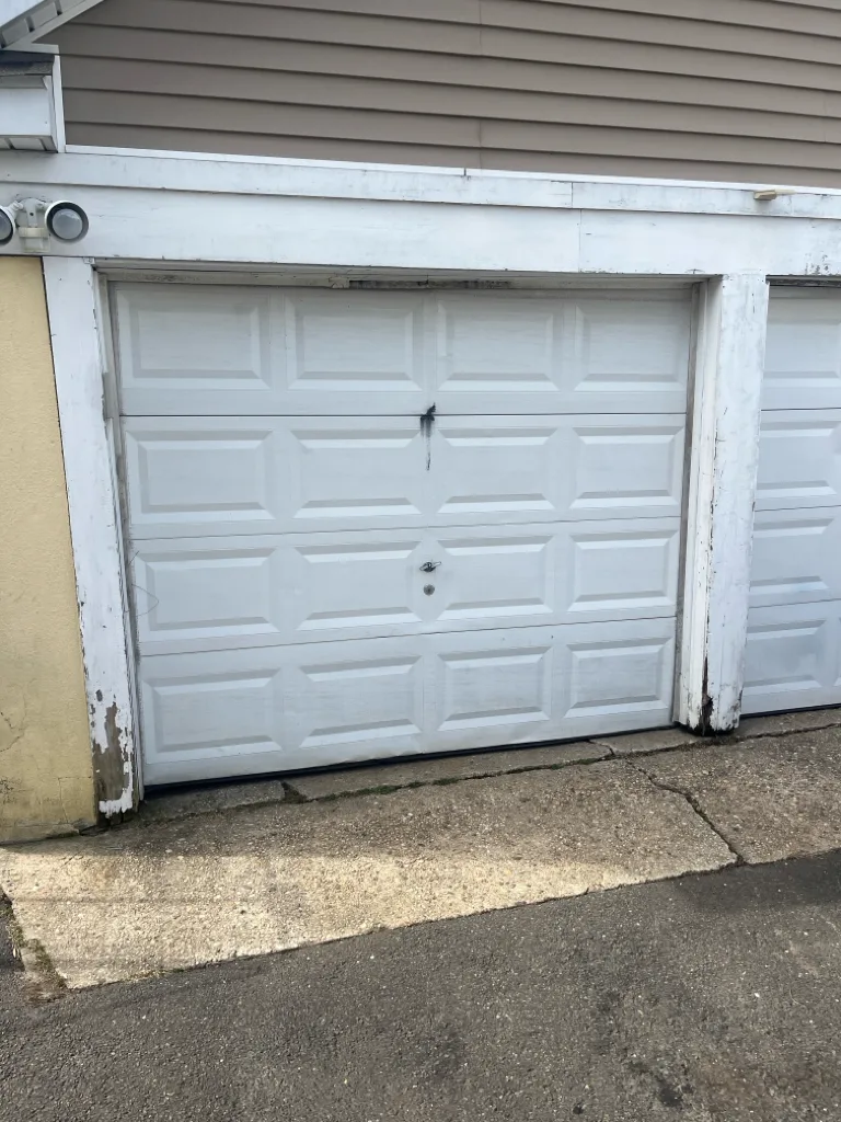 A white raised-panel garage door in a wooden opening whose jamb and trim have blistered and split at the base, the paint peeling away above a stained concrete apron.