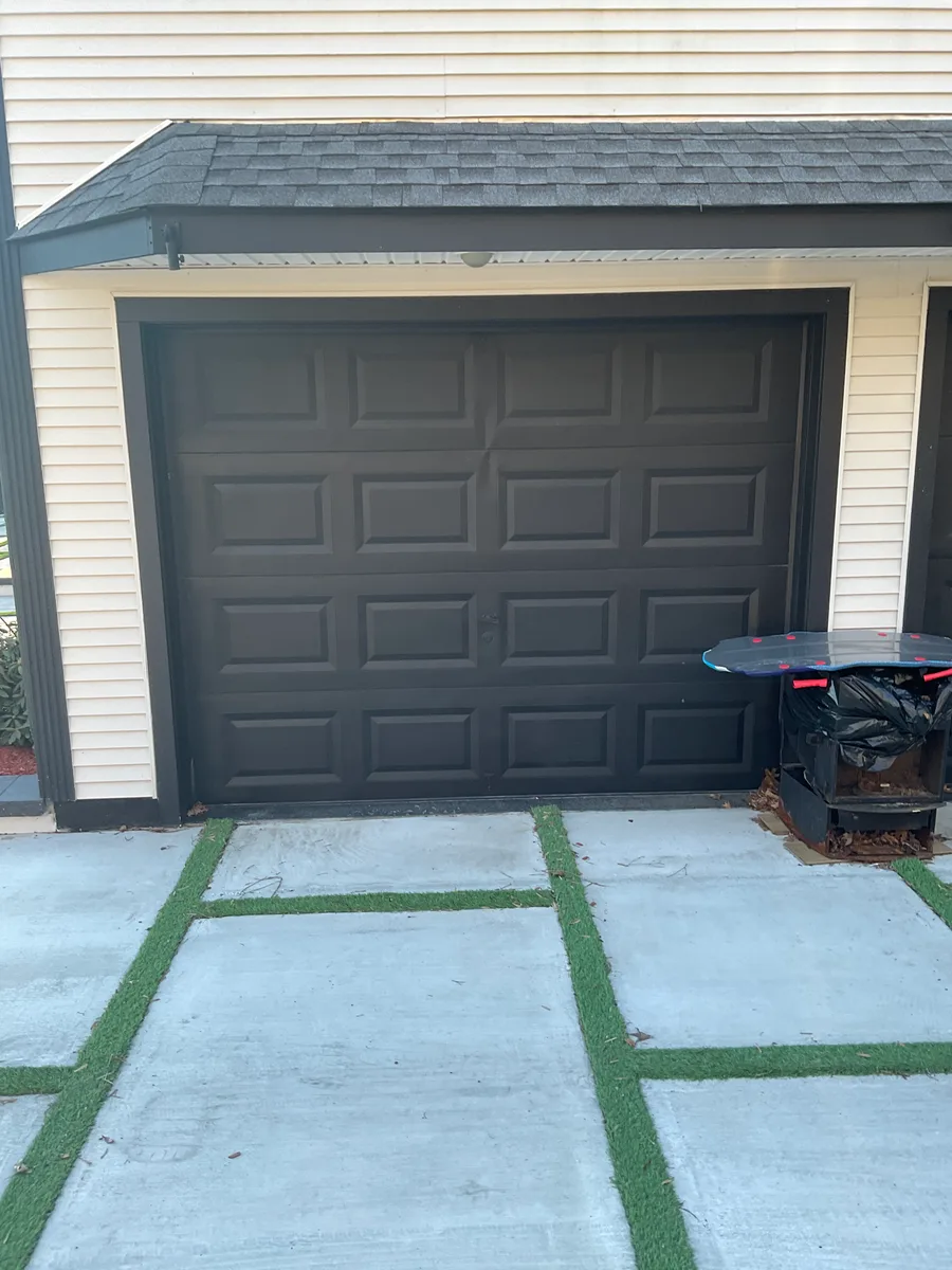 A dark bronze raised-panel garage door under its own shingled roof overhang on a cream-sided house, with a paver-and-turf apron in front of it.