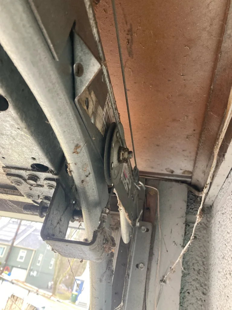 A grubby cable drum and end bearing plate at the top corner of a garage door, the lift cable rising off the drum with the curved track and roller carriage beside it.