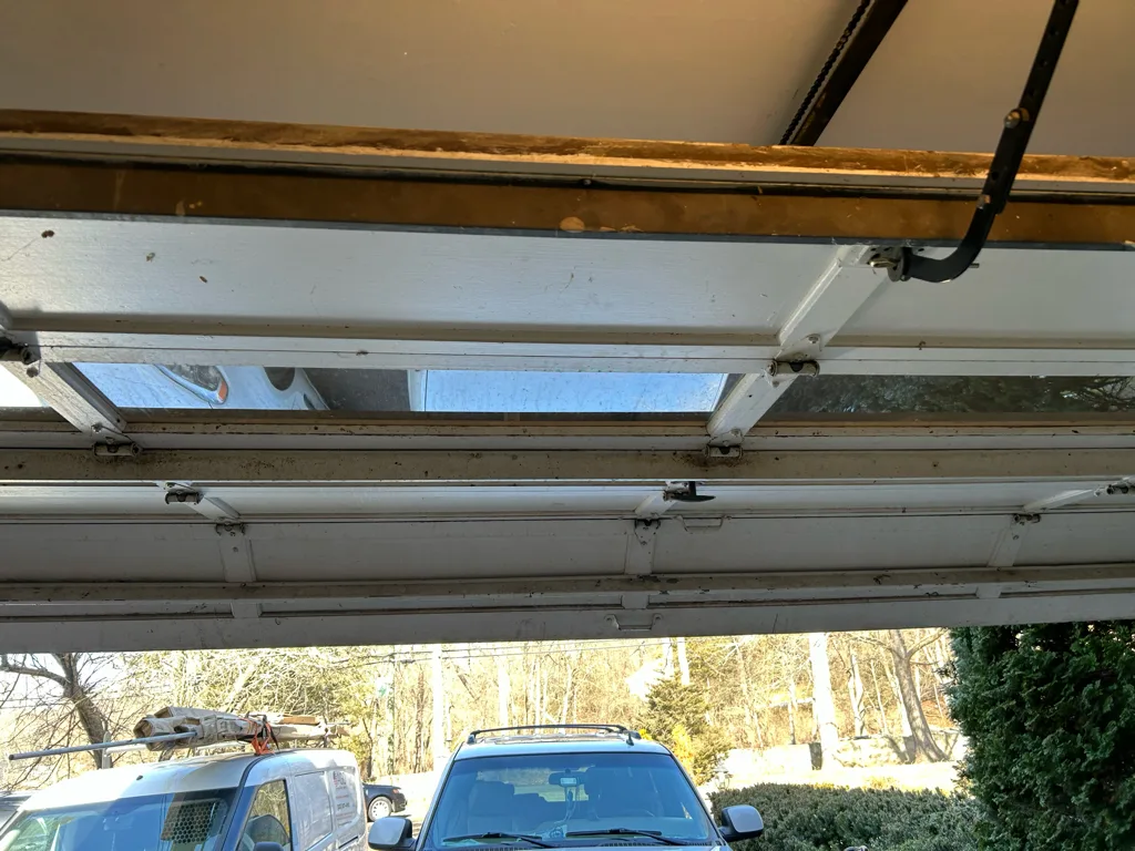 Looking up from inside at a partly raised garage door, sunlight coming through the glazed section and catching the hinges and roller stems along the joint between panels.