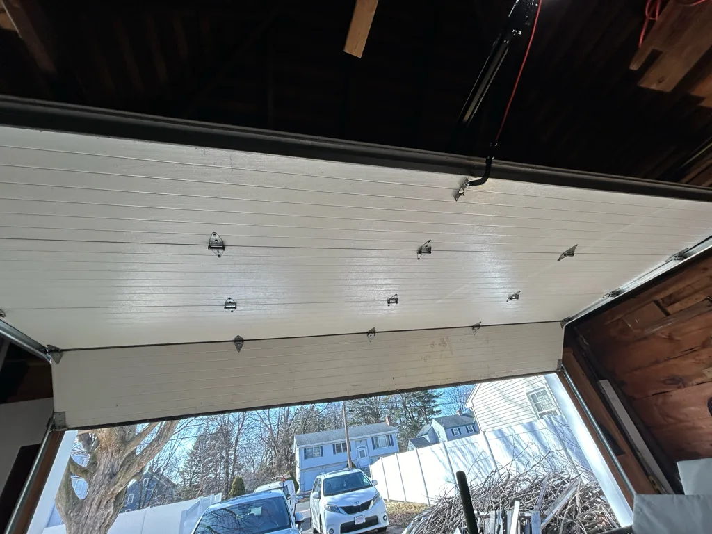 The underside of a garage door raised into its horizontal tracks, hinges and roller brackets running across the panels, with a neighbouring house and parked cars visible through the open bay.
