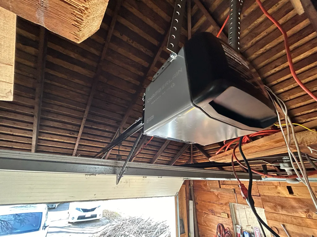 A chain-drive opener bolted up into the exposed rafters of a garage, its rail angling down to the door and mains and sensor wiring looped along the timbers beside it.